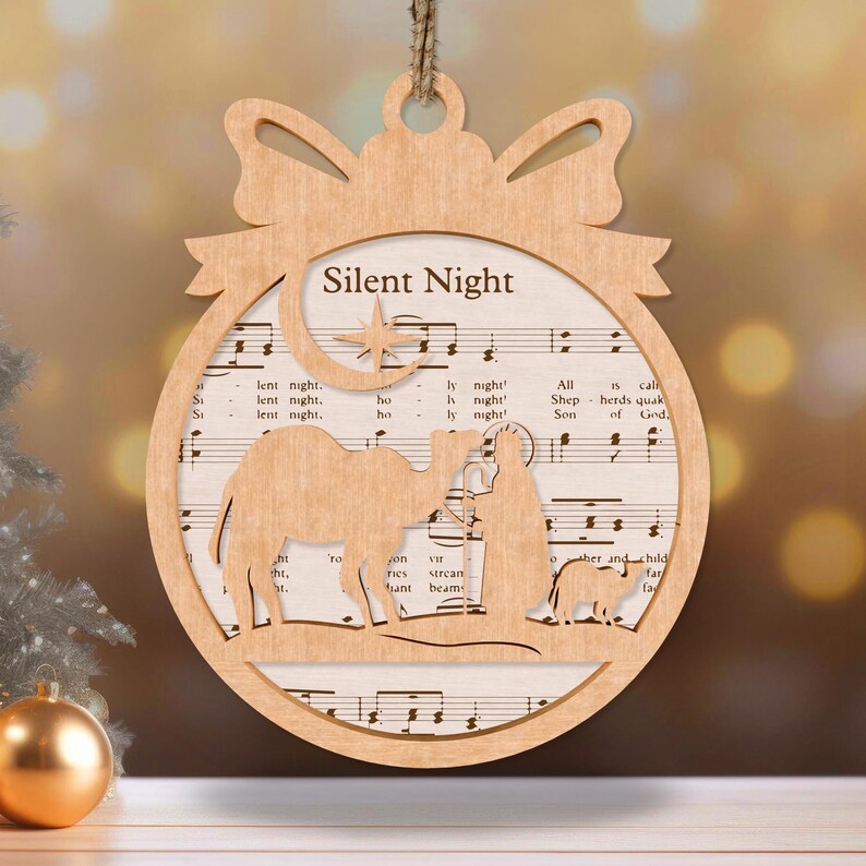May include: A wooden Christmas ornament shaped like a circle with a bow at the top. The ornament features a cutout of the nativity scene with a star and a crescent moon. The ornament is decorated with sheet music for the song "Silent Night".