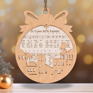 May include: A wooden Christmas ornament shaped like a circle with a bow at the top. The ornament is decorated with a cutout of the nativity scene and the lyrics to the Christmas carol "O, Come All Ye Faithful".