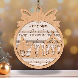 May include: A wooden Christmas ornament shaped like a circle with a bow at the top. The ornament features a cutout of the nativity scene with the three wise men and their camels. The ornament is decorated with the sheet music for the song "O Holy Night".