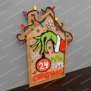 May include: Wooden Christmas countdown sign featuring a Grinch hand, snowflakes, and colorful lights. The sign reads "24 Days Until Christmas" in orange and white lettering. The house-shaped sign is made of wood.