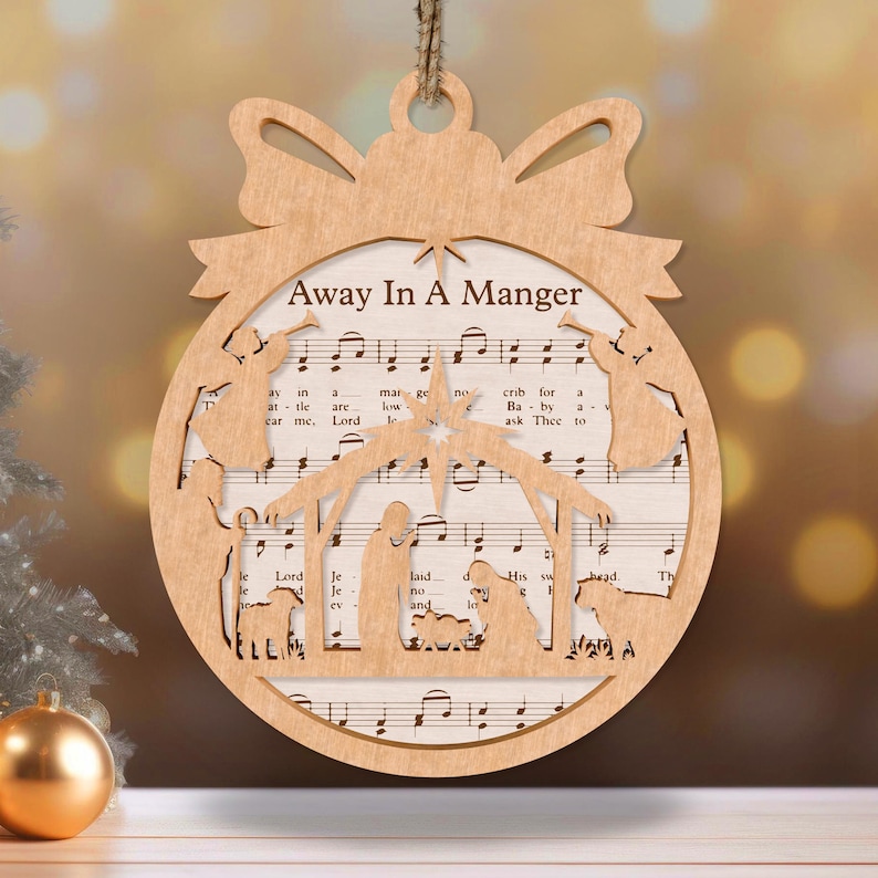 May include: A wooden Christmas ornament shaped like a circle with a cutout of the nativity scene. The ornament is decorated with the words "Away In A Manger" and the sheet music for the song.