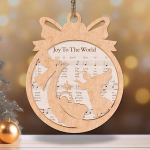 May include: A wooden Christmas ornament shaped like a ball with a bow at the top. The ornament is decorated with a silhouette of angels and the words "Joy To The World".