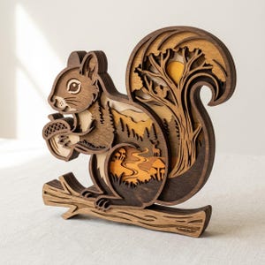 May include: A multi-layered wooden squirrel sculpture featuring a dark brown squirrel holding an acorn while perched on a branch. The interior cutout reveals a forest scene with trees, a deer, mushrooms, and a sun in shades of tan and orange.