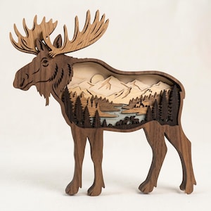 May include: A multi-layered wooden moose silhouette featuring a cut-out landscape scene. The interior displays a mountain range, a winding river, and a dense forest of pine trees with small moose figures, all crafted from various shades of wood.
