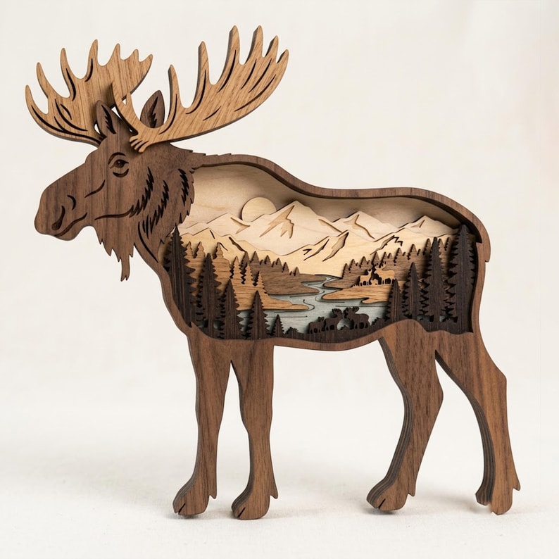 May include: A multi-layered wooden moose silhouette featuring a cut-out landscape scene. The interior displays a mountain range, a winding river, and a dense forest of pine trees with small moose figures, all crafted from various shades of wood.