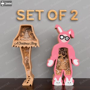 May include: A set of two laser-cut wooden decorations, 3mm thick. One is a leg lamp shape with the text A Christmas Story, and the other is a pink bunny suit figure with black glasses. Both feature intricate layered cutouts of holiday scenes.