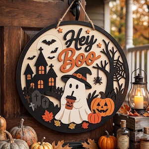 Puede incluir: Un letrero de puerta redondo de madera para Halloween con el texto Hey Boo en negro y naranja. El diseño incluye un fantasma blanco con sombrero de bruja, una casa encantada negra, calabazas naranjas, murciélagos y hojas de otoño.