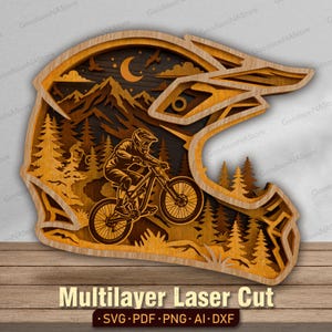 May include: A multilayered laser-cut wooden wall art piece shaped like a motocross helmet. The interior features a detailed scene of a mountain biker riding through a forest with mountains, a crescent moon, and birds in the background.