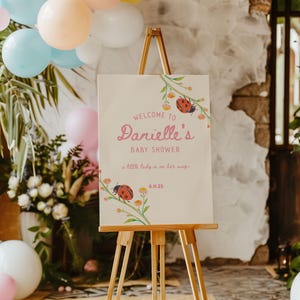 May include: A wooden easel displays a baby shower sign. The sign reads "Welcome to Danielle's Baby Shower" in pink script, with ladybug and floral accents. The date 6.11.25 is at the bottom. Balloons and floral arrangements are in the background.
