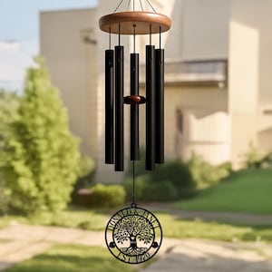 Op de afbeelding: Een windgong met vijf matzwarte cilindrische metalen buizen die aan een ronde houten bovenkant hangen. Een centrale houten klepel hangt tussen de buizen. Een ronde zwarte metalen hanger toont een boomontwerp met de tekst DONALD & JANET EST. 1970.