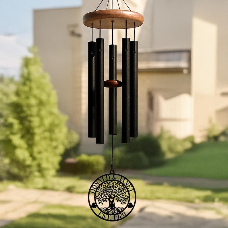 Op de afbeelding: Een windgong met vijf matzwarte cilindrische metalen buizen die aan een ronde houten bovenkant hangen. Een centrale houten klepel hangt tussen de buizen. Een ronde zwarte metalen hanger toont een boomontwerp met de tekst DONALD & JANET EST. 1970.