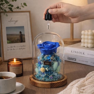 May include: A preserved blue rose encased in a clear glass dome on a circular wooden base. The arrangement includes light blue and white dried flowers and greenery. A small black dropper is positioned above the dome, and a rectangular tag with the text Clarre is attached to the floral base.