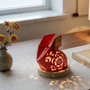 Könnte beinhalten: Rote Keramiklampe in Hasenform mit floralem Design, auf einem Holzsockel. Die Lampe ist beleuchtet und strahlt ein warmes Licht aus. Die Ohren haben goldene Akzente. Die Lampe steht auf einem Tisch.