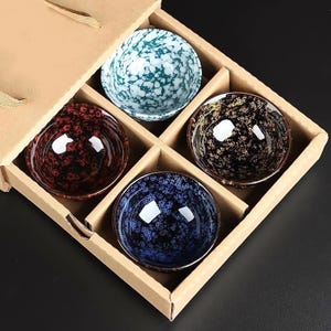 May include: A set of four ceramic bowls in a cardboard gift box. Each bowl features a unique speckled pattern in teal, red, gold, and blue. The bowls have a glossy finish and are designed for serving. The box has a natural brown color.