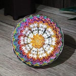 May include: A decorative, iridescent dish with a floral design. The dish features a central gold flower surrounded by white and peacock feather-like patterns, with a rainbow-colored rim. The dish is on a wooden surface.