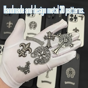 Puede incluir: Una mano enguantada de blanco sosteniendo varios patrones 3D de metal plateado, incluyendo cruces, flores de lis y una herradura con una cruz en el centro. El texto "Handmade and design metal 3D patterns." est&aacute; en la parte superior de la imagen.