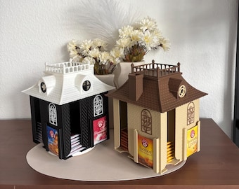 May include: Two house-shaped tea bag organizers with rotating bases. One unit is black with a white roof, and the other is beige with a brown roof. Both feature arched windows, a rooftop railing, and slots to hold tea bags with visible labels.