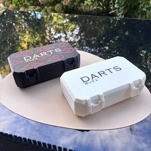 Könnte beinhalten: Ein schwarzes rechteckiges Dart-Etui mit roten Wolkenmustern und dem Text DARTS sowie ein weißes rechteckiges Etui mit dem Text DARTS. Beide Etuis haben einen Klappdeckel mit Verschluss und japanische Schriftzeichen unter dem Wort DARTS.