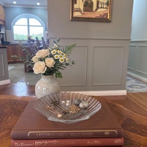 May include: A clear, scalloped glass dish holds a vintage-style key, two pearl earrings, two smooth grey stones, and gold-toned jewellery. The dish rests on two stacked hardcover books titled A Victorian Family Molly Hughes and The Illustrated Brontës.