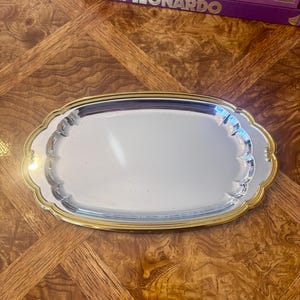 May include: An oval serving tray with a polished silver-tone centre and a decorative gold-tone beaded rim. The tray features a scalloped edge design with small ornate details at the sides, providing a reflective surface for serving or display.
