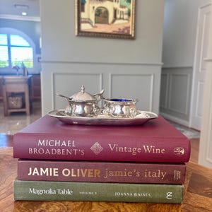 May include: A silver condiment set with a lidded pot and an open bowl on an oval tray, resting atop three stacked hardcover books titled Michael Broadbent's Vintage Wine, Jamie Oliver's Jamie's Italy, and Joanna Gaines's Magnolia Table Volume 3.