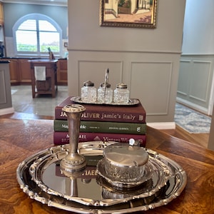 May include: A silver-plated serving tray holding a tall, slender bud vase with a decorative base and a small glass condiment jar with a silver lid. Behind these, a stack of three books supports a silver caddy containing three small glass shakers.