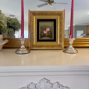 May include: A small oil painting in a thick, ornate gold frame with a black inner border, depicting a floral arrangement in a vase. The painting is flanked by two silver-toned candle holders, each containing a tall, spiralled, metallic pink candle.
