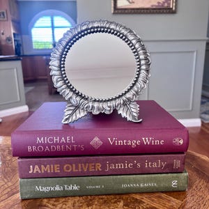 May include: A round silver-toned metal mirror with a decorative beaded and leaf-patterned frame, resting on a stack of three books titled Michael Broadbent's Vintage Wine, Jamie Oliver's Jamie's Italy, and Joanna Gaines's Magnolia Table Volume 3.