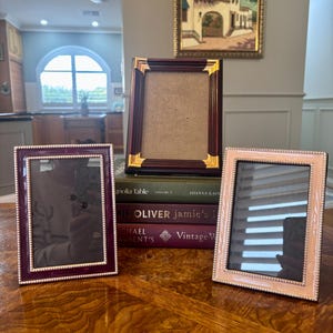 May include: Three decorative picture frames in burgundy, gold, and light pink. The frames are displayed on a wooden table, with a painting in the background. The frames have ornate detailing and are empty, ready for photos.