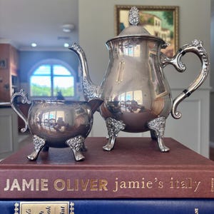 May include: A silver tea set, comprising a teapot and sugar bowl with intricate patterns, rests on two cookbooks. The teapot features a curved spout and a decorative handle. The sugar bowl has a matching design. The books are titled "Jamie Oliver" and "Gourmet's Basic French Cookbook."