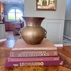 May include: A brass vase with a flared rim and side ring handles sits atop a stack of four books. The books are titled Oil Painting Pure and Simple, A Standard Guide to Pure-Bred Dogs, The Complete Guide to Flower Arranging, and Leonardo.
