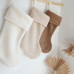 Peut inclure: Trois bas de Noël en peluche crème, beige et marron sont suspendus à une tringle en bois contre un mur de briques blanches. Chaque bas a un revers plié et une boucle pour l'accrocher. Les bas sont en matière douce et texturée.