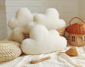 May include: A collection of cloud-shaped pillows, a woven basket, and a mushroom-shaped basket. The pillows are white and fluffy. A wooden brush and a woven rattle are also present. The items are arranged on a white cloth.