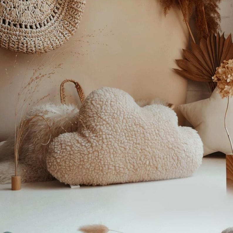 May include: A cream-coloured, cloud-shaped cushion with a soft, textured surface. The cushion is part of a collection of decorative items, including a star-shaped cushion and woven wall decor. The cushion is approximately 40 cm in length.