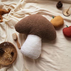 May include: A brown and white plush mushroom shaped pillow. The mushroom is on a white textured fabric background.