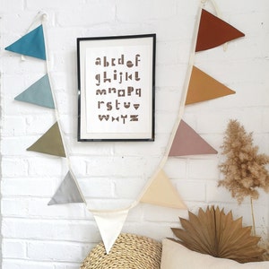 May include: A white string of triangle flags with brown, beige, light blue, grey, and pink fabric. The flags are hanging on a white brick wall. A framed print with the alphabet in brown letters is on the wall.