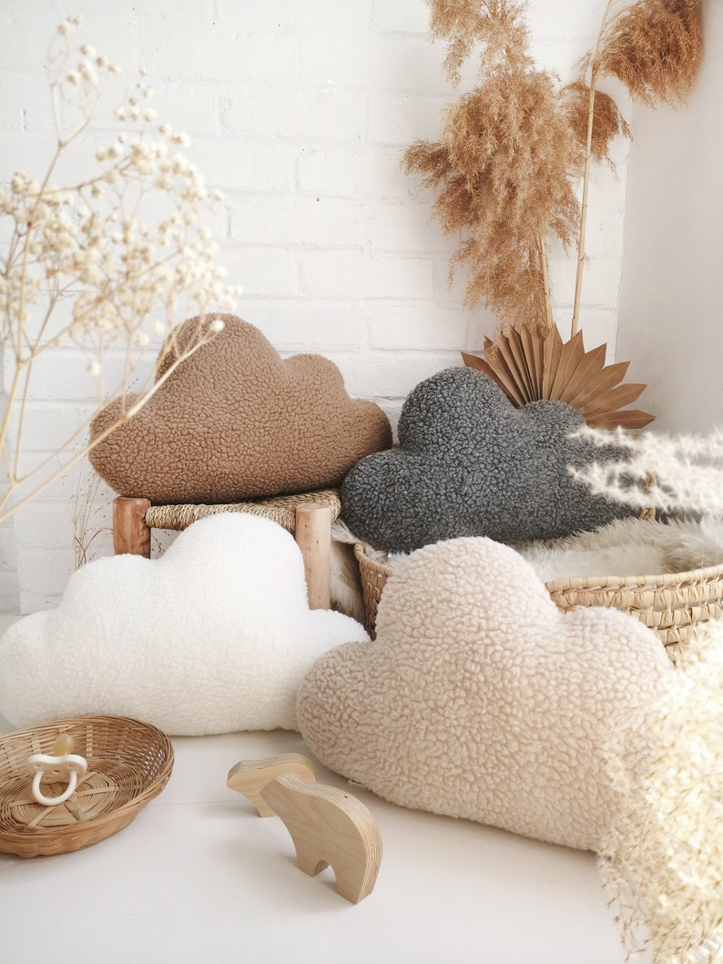 May include: Four cloud-shaped cushions in shades of brown, grey, white, and beige. The cushions have a fluffy, textured appearance. A wooden bear toy and a small woven basket with a dummy are in the foreground.