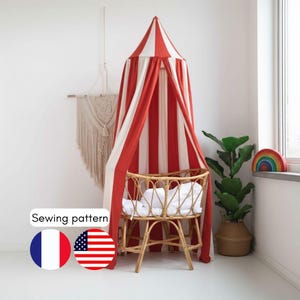 Puede incluir: Un dosel a rayas rojas y blancas cuelga sobre una cuna de mimbre con un colchón blanco. Un tapiz de macramé y una planta en maceta también están en la habitación. La imagen incluye el texto "Sewing pattern" y las banderas de Francia y Estados Unidos.