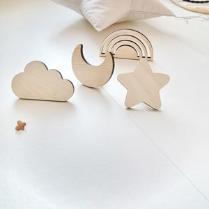 Puede incluir: Formas de madera recortadas de una nube, una luna creciente, un arcoíris y una estrella. Las formas son de color marrón claro y tienen un acabado de madera natural.