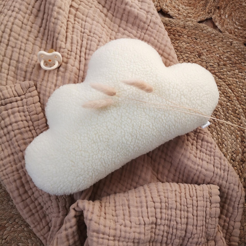May include: A soft, white cloud-shaped cushion with a textured surface. The cushion is decorated with a few sprigs of dried, pale pink grass. A dummy is visible in the upper left corner. The cushion rests on a pink blanket.