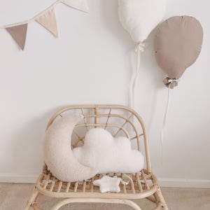 May include: A nursery scene with a rattan chair, moon and cloud cushions, and balloon-shaped pillows. A banner of triangular flags hangs on the wall. The colour scheme is neutral, with beige and white tones. The chair is made of natural rattan.