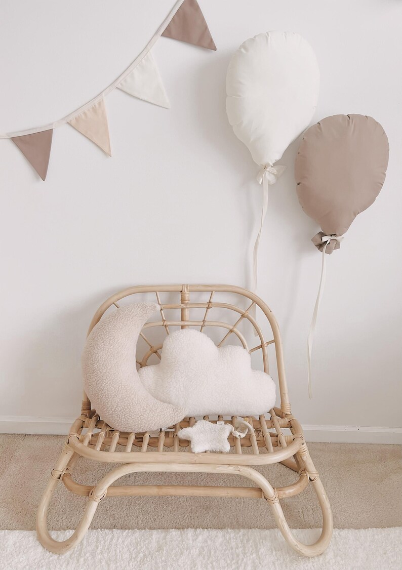 May include: A nursery scene with a rattan chair, moon and cloud cushions, and balloon-shaped pillows. A banner of triangular flags hangs on the wall. The colour scheme is neutral, with beige and white tones. The chair is made of natural rattan.