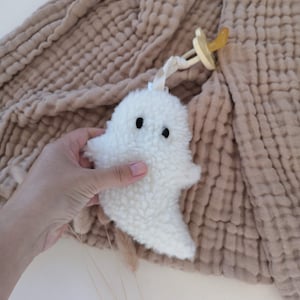 May include: A white plush ghost-shaped baby toy with black button eyes and a pacifier attached. The toy is made of a soft, textured material. The background features a beige, crinkled fabric.