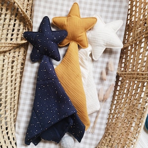 May include: Three star-shaped muslin baby swaddles in a wicker basket. One swaddle is navy blue with gold glitter, one is mustard yellow, and one is white.