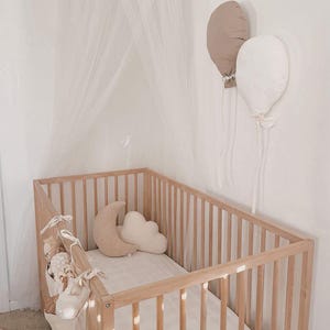 May include: A light wood baby cot with a white mattress, featuring beige moon and cloud-shaped pillows. Two balloon-shaped cushions, one beige and one white, are on the wall. A sheer canopy hangs above the cot, creating a soft, neutral nursery setting.