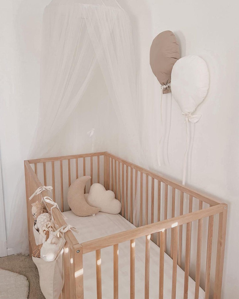 May include: A light wood baby cot with a white mattress, featuring beige moon and cloud-shaped pillows. Two balloon-shaped cushions, one beige and one white, are on the wall. A sheer canopy hangs above the cot, creating a soft, neutral nursery setting.