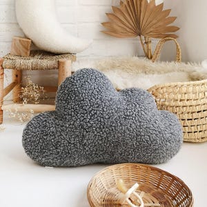 May include: A grey cloud-shaped cushion made from a soft, textured fabric. The cushion is the main focus, set against a neutral backdrop with a wicker basket, a small wooden stool, and a crescent-shaped pillow.