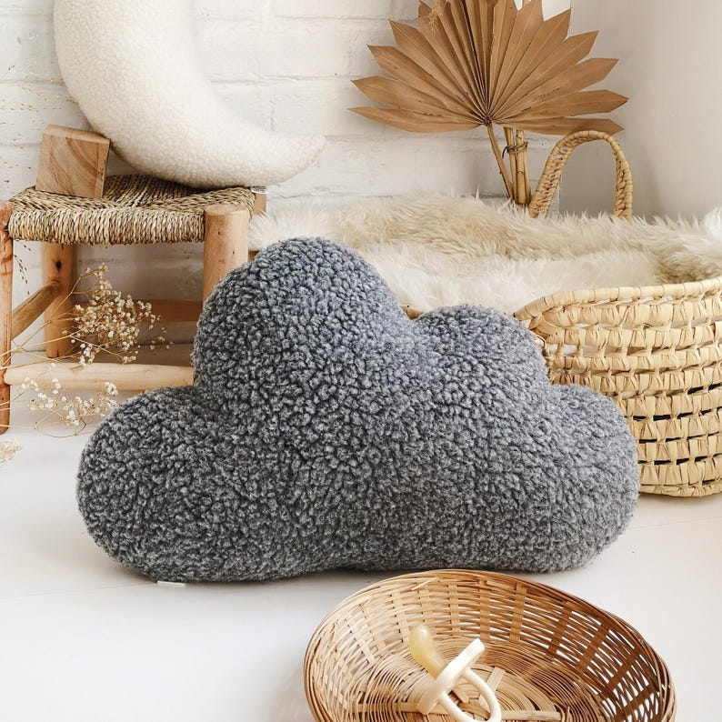May include: A grey cloud-shaped cushion made from a soft, textured fabric. The cushion is the main focus, set against a neutral backdrop with a wicker basket, a small wooden stool, and a crescent-shaped pillow.