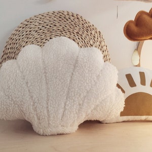 Puede incluir: Una almohada de concha blanca de felpa con respaldo de mimbre tejido. La almohada está sobre una superficie de madera junto a una almohada de sol de color tostado con contorno blanco.