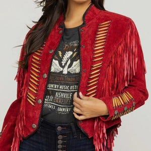 May include: Red suede jacket with fringe and decorative beading. The jacket has a round collar and metal buttons. A black graphic t-shirt is visible underneath the jacket. The t-shirt has text that says "Nashville Country Music Festival".